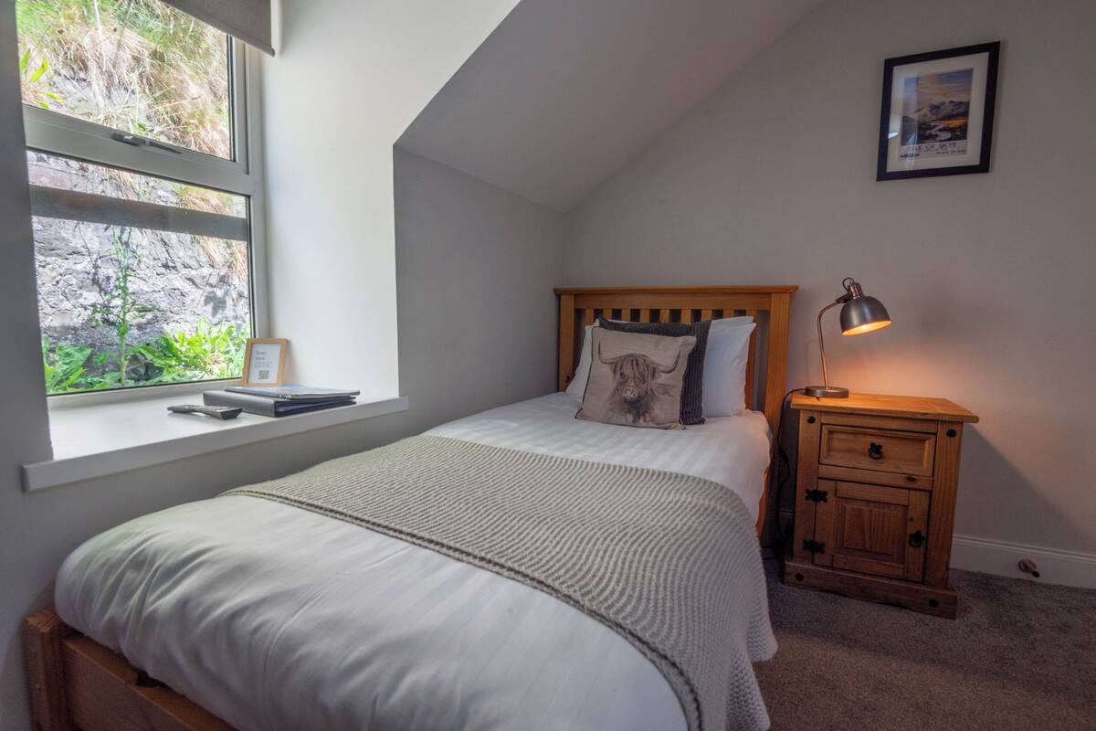 A bedroom with a bed, a nightstand with a lamp, a window with a view of greenery, and a framed picture on the wall.