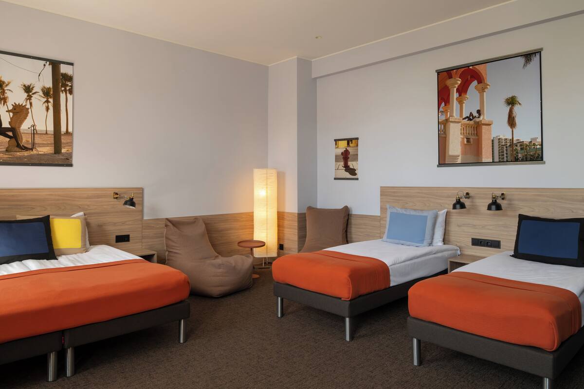 Ett hotellrum med tre sängar, en sittpuff, ett litet bord och en vägglampa.