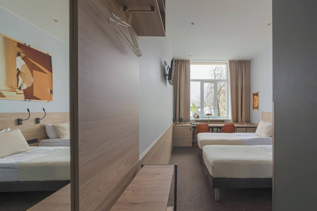 Ett hotellrum med två sängar, ett skrivbord, en stol, ett fönster med gardiner och en TV som är monterad på väggen.