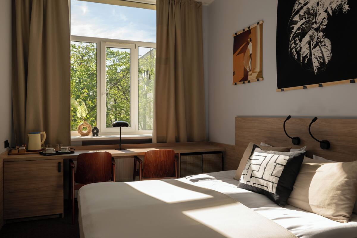 Ett hotellrum med en säng, ett skrivbord, en stol, ett fönster med gardiner och konstverk på väggarna.