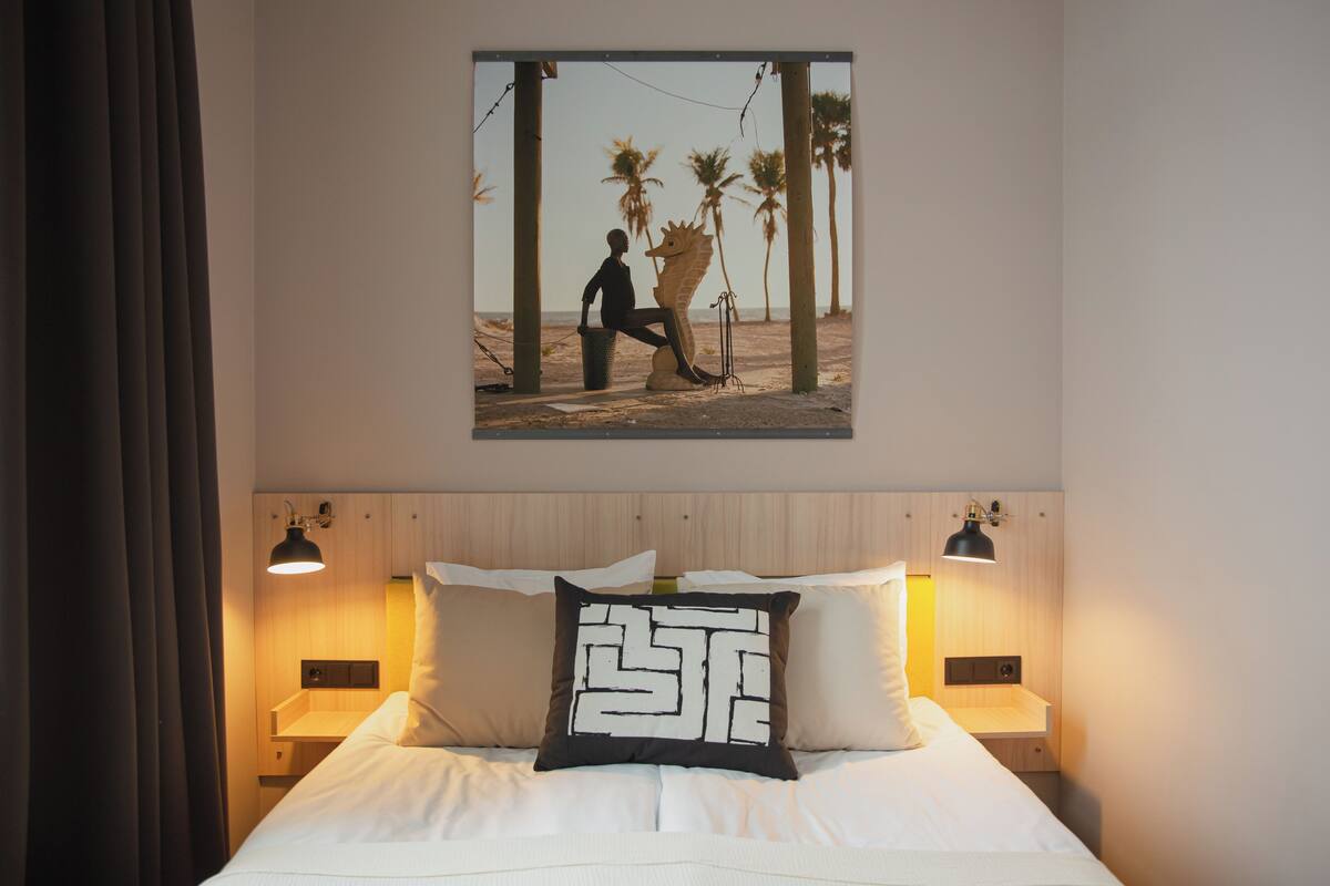 Ett hotellrum med en säng, ett nattduksbord, en tavla som hänger på väggen och en kudde med en unik design.