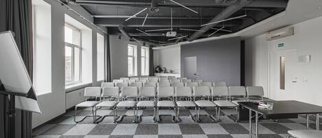 Un salón de conferencias con piso a cuadros, filas de sillas blancas, un proyector y una pizarra.
