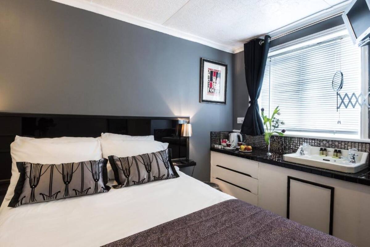 A hotel room with a bed, a nightstand, a lamp, a window with curtains, a sink, and a framed picture on the wall.