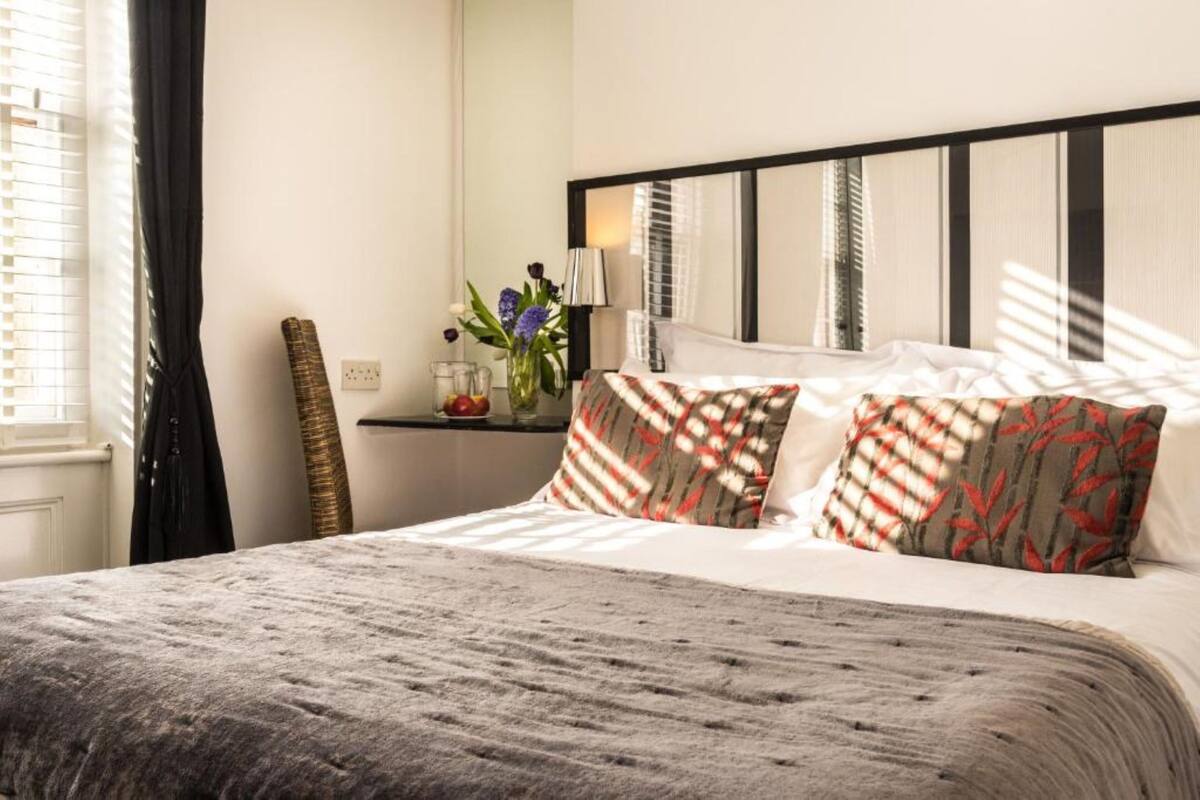 A neatly made bed with patterned pillows, a bedside table with a vase of flowers, and a window with blinds.