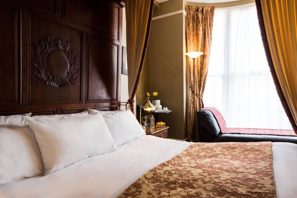 A hotel room with a large bed, a wooden headboard, a nightstand with a lamp, and a view of the outdoors through curtains.