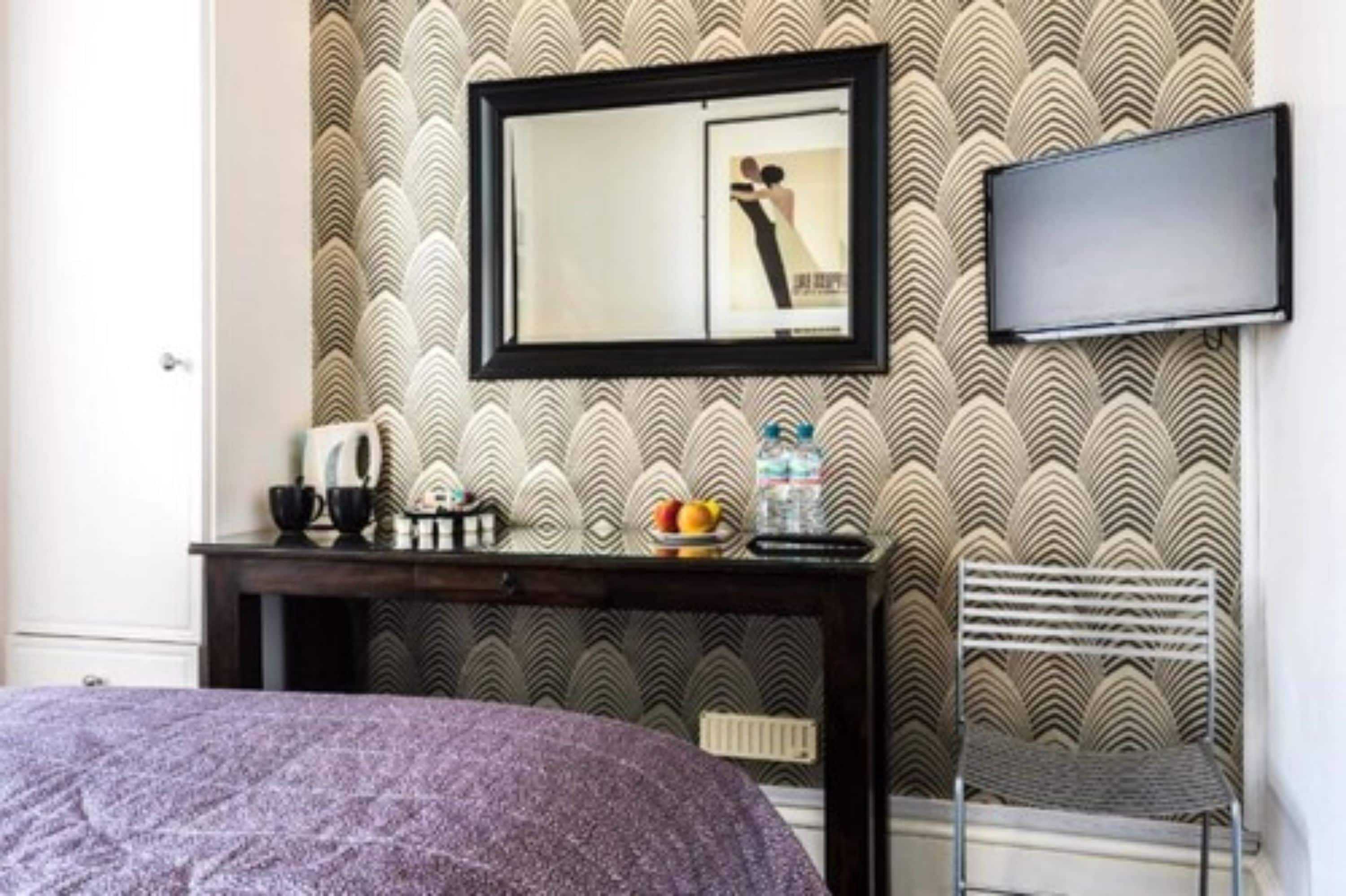A hotel room with a bed, a desk with a mirror, a television, and a patterned wallpaper.