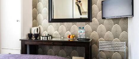 A hotel room with a bed, a desk with a mirror, a television, and a patterned wallpaper.