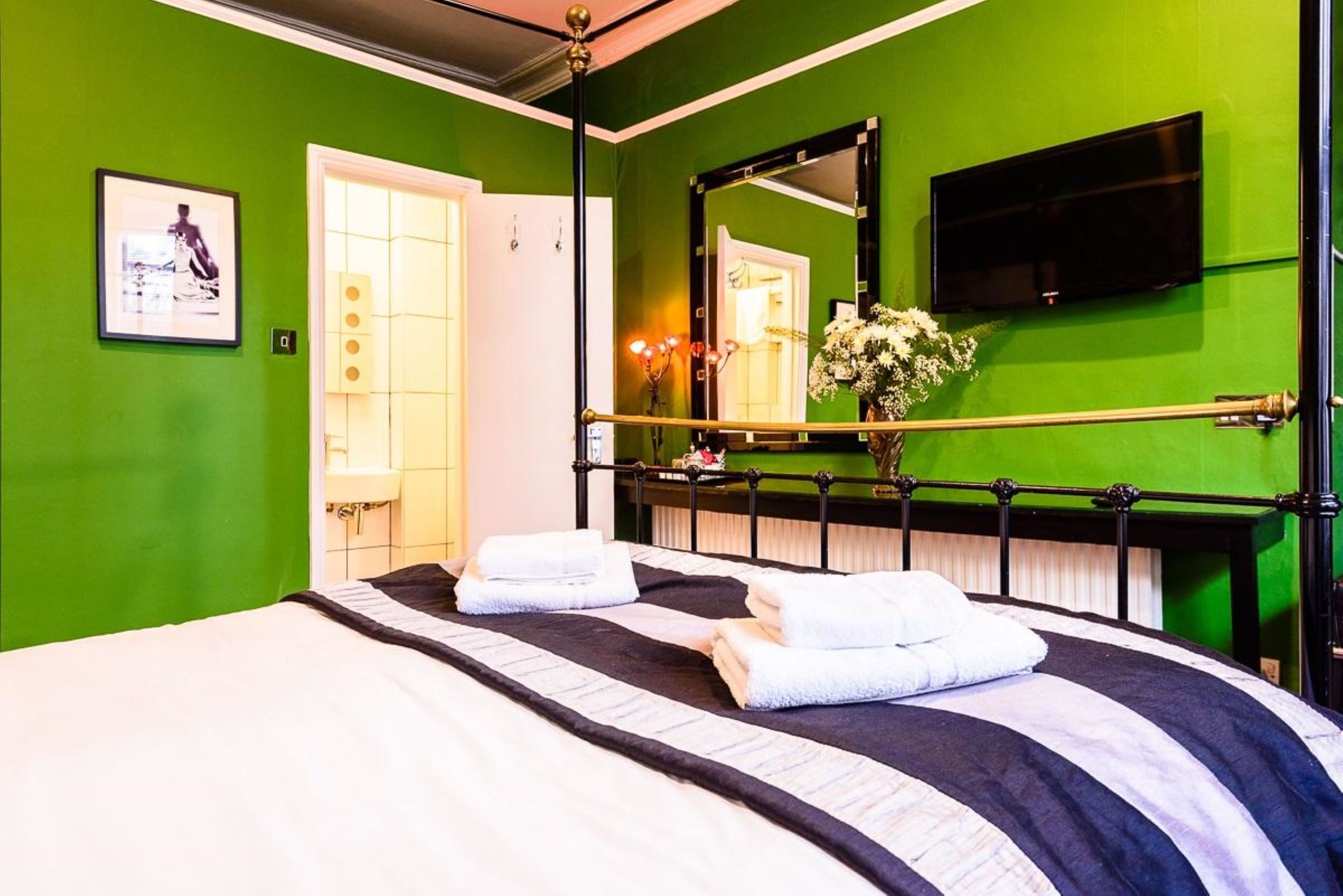 A hotel room with a green wall, a bed with white and black bedding, a black metal headboard, a flat-screen TV mounted on the wall, and a mirror above the bed.