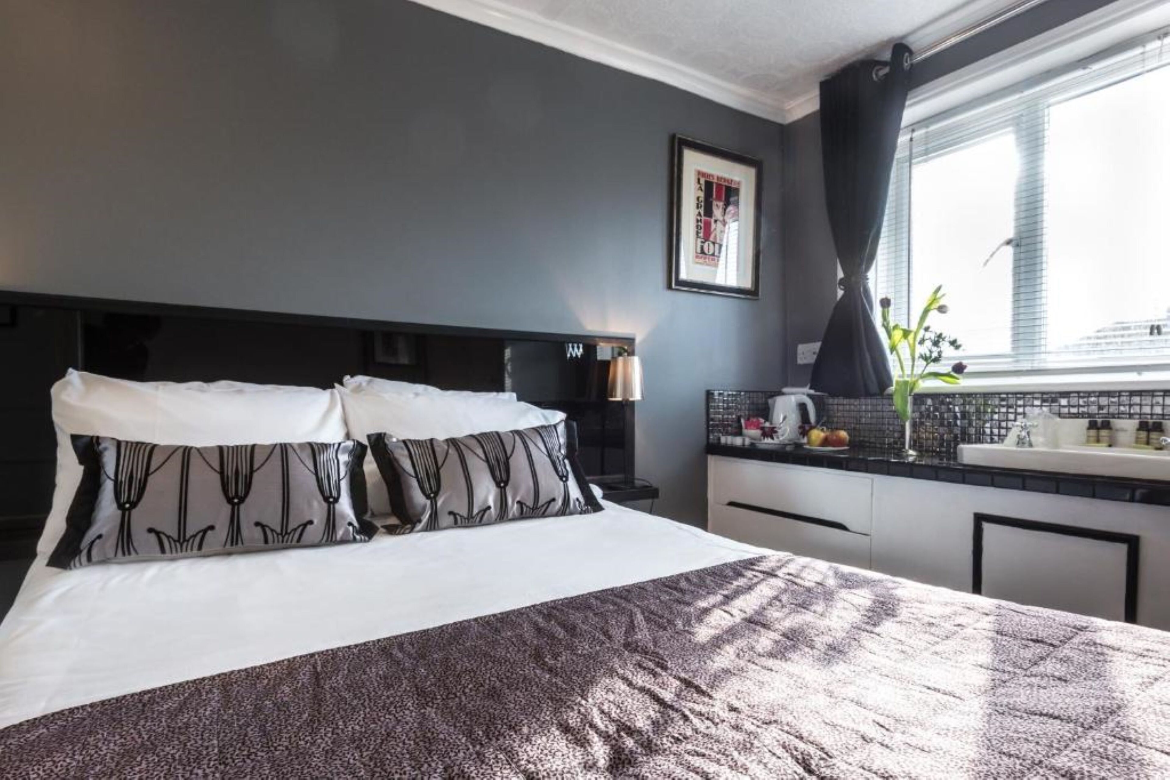 Una habitación de hotel con cama, mesita de noche, ventana con cortinas y una zona de kitchenette.