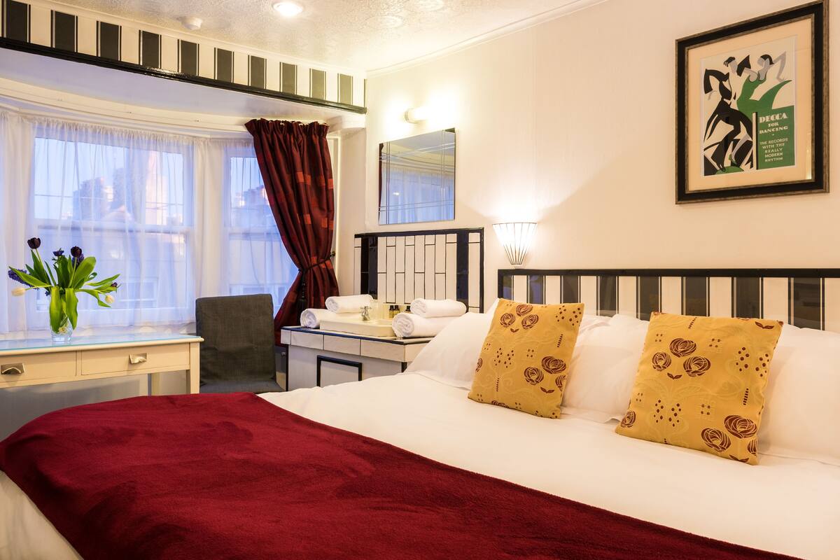 A hotel room with a large bed, a desk with a chair, a window with curtains, a mirror, and a framed poster on the wall.