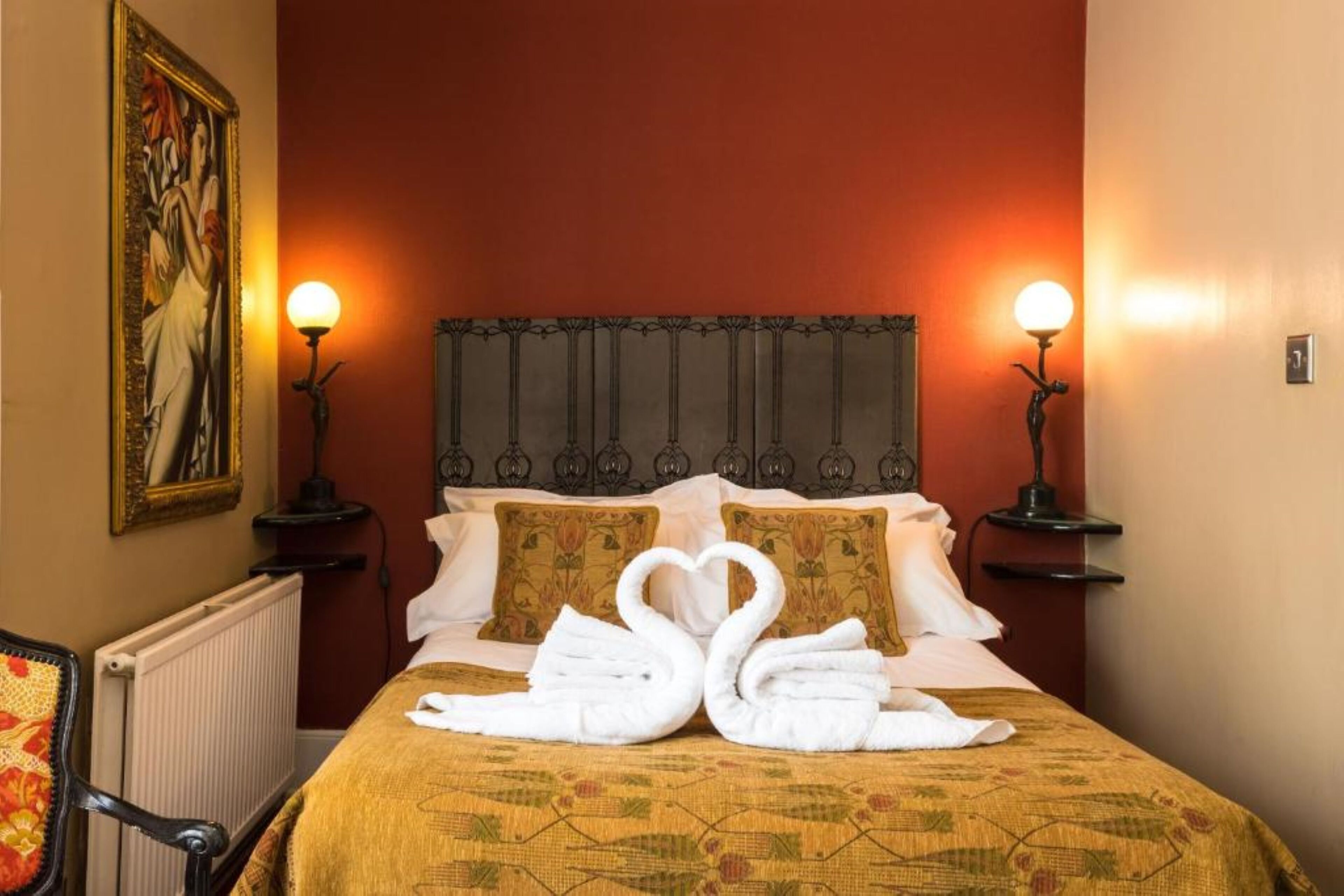 A hotel room with a bed, two wall-mounted lamps, a radiator, and a painting on the wall.