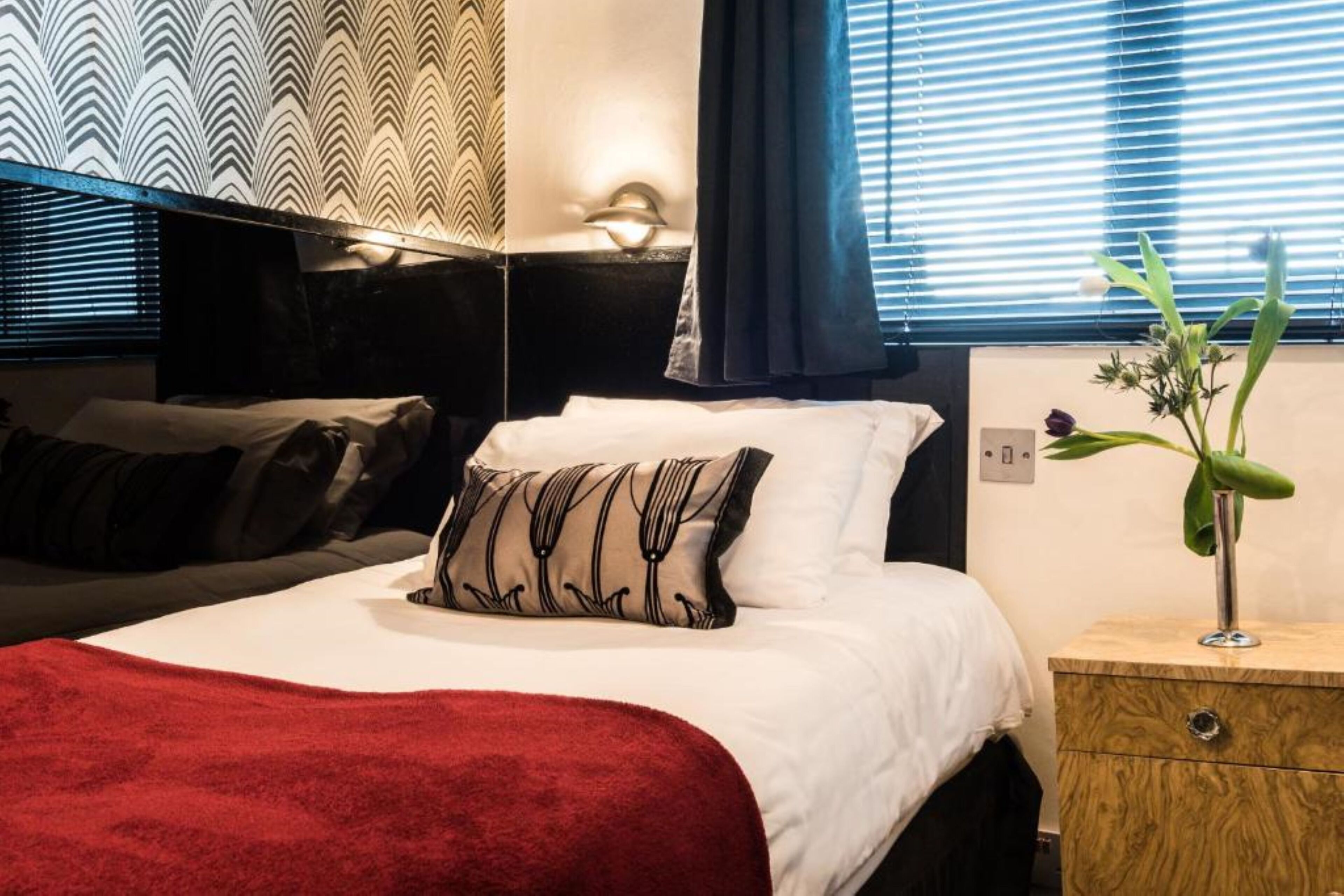 A neatly made bed with a red blanket, a bedside table with a vase of flowers, and a patterned wallpaper.