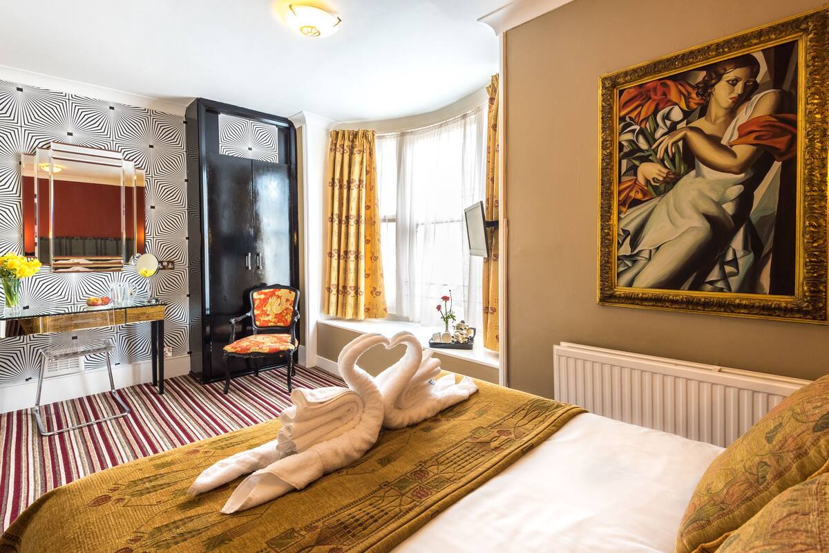 A hotel room with a bed, a desk with a mirror, a chair, a window with curtains, and a painting on the wall.