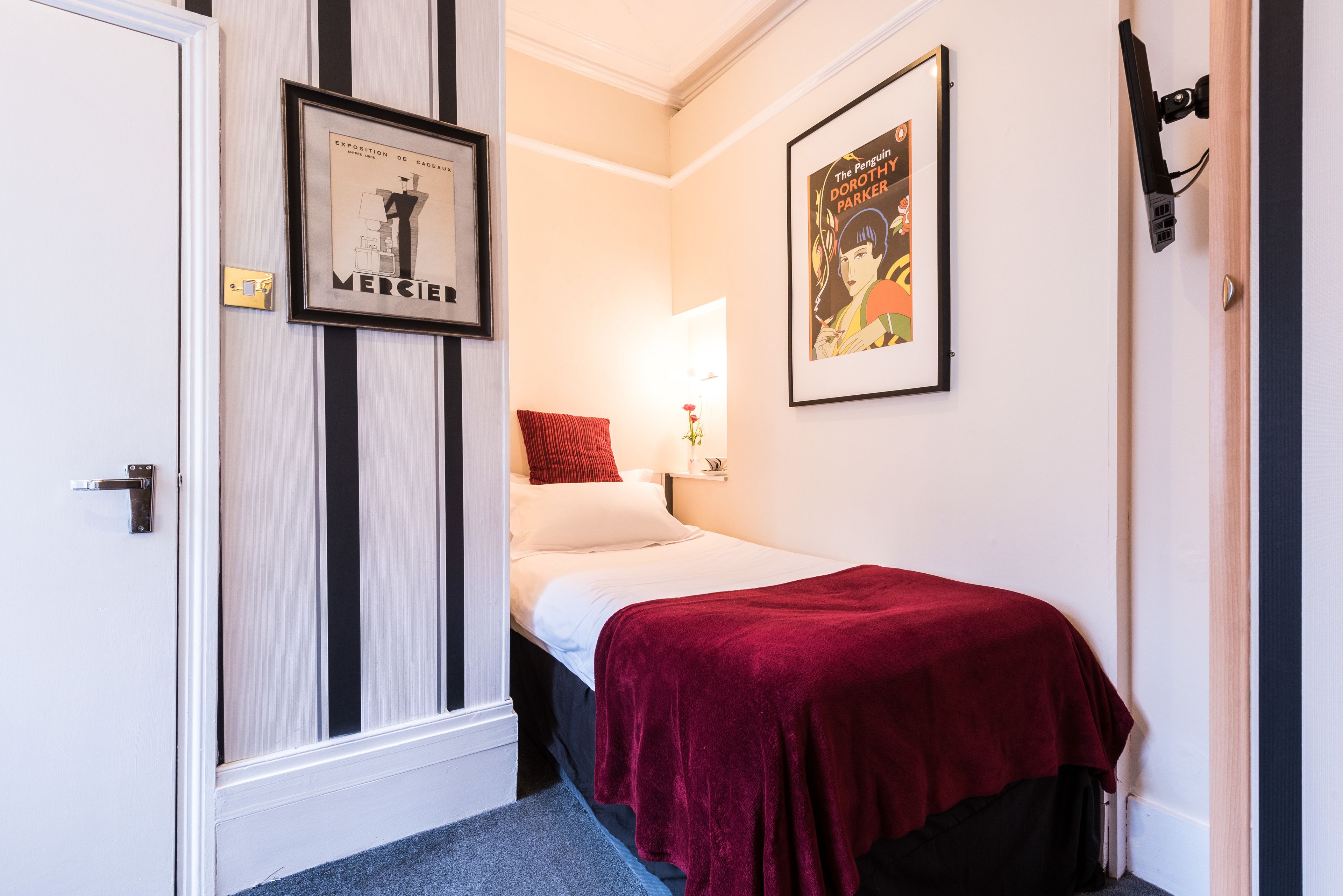 A hotel room with a bed, a red throw blanket, a framed picture on the wall, and a door with a handle.