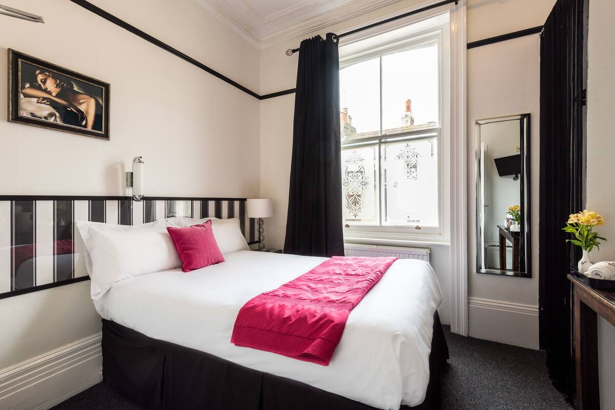 A hotel room with a large bed, a window with curtains, a bedside table with a lamp, and a framed picture on the wall.