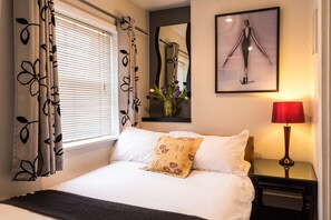 Un lit bien fait, avec du linge de lit blanc et un coussin décoratif, une table de chevet avec une lampe rouge et une œuvre d’art encadrée au-dessus du lit.