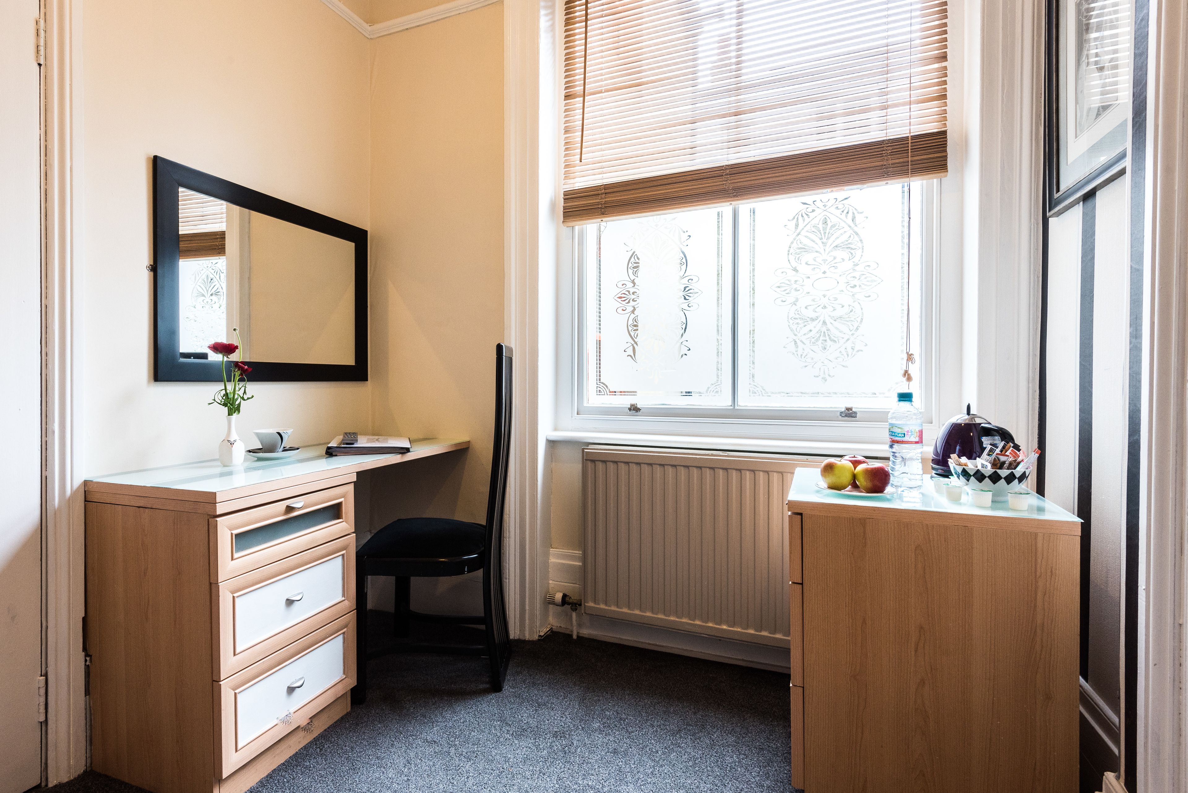 A bedroom with a bed, a bedside table, a dresser, a window with blinds, and a mirror.