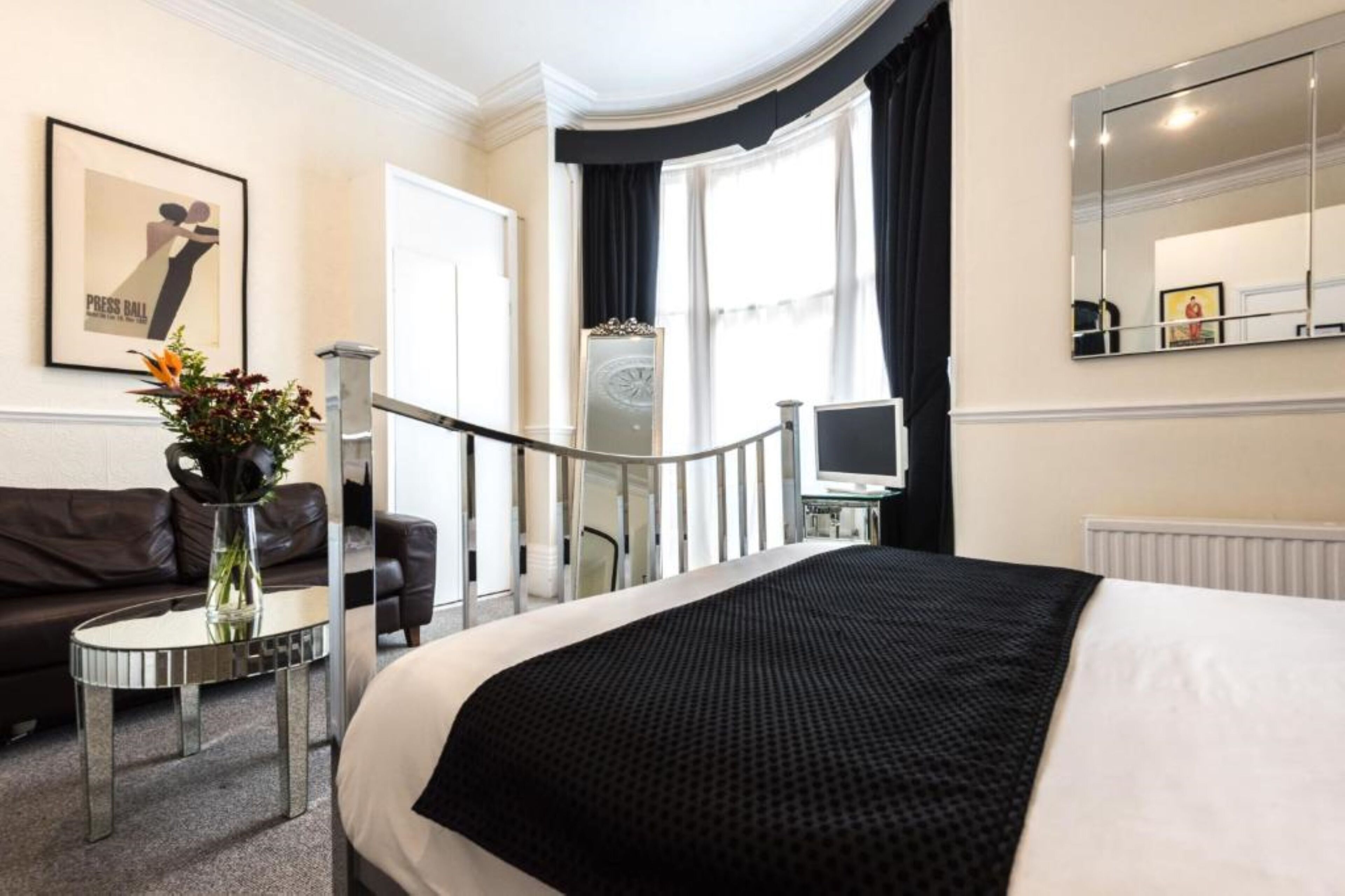A hotel room with a large bed, a small round table with a vase of flowers, a sofa, a television, and a large mirror.
