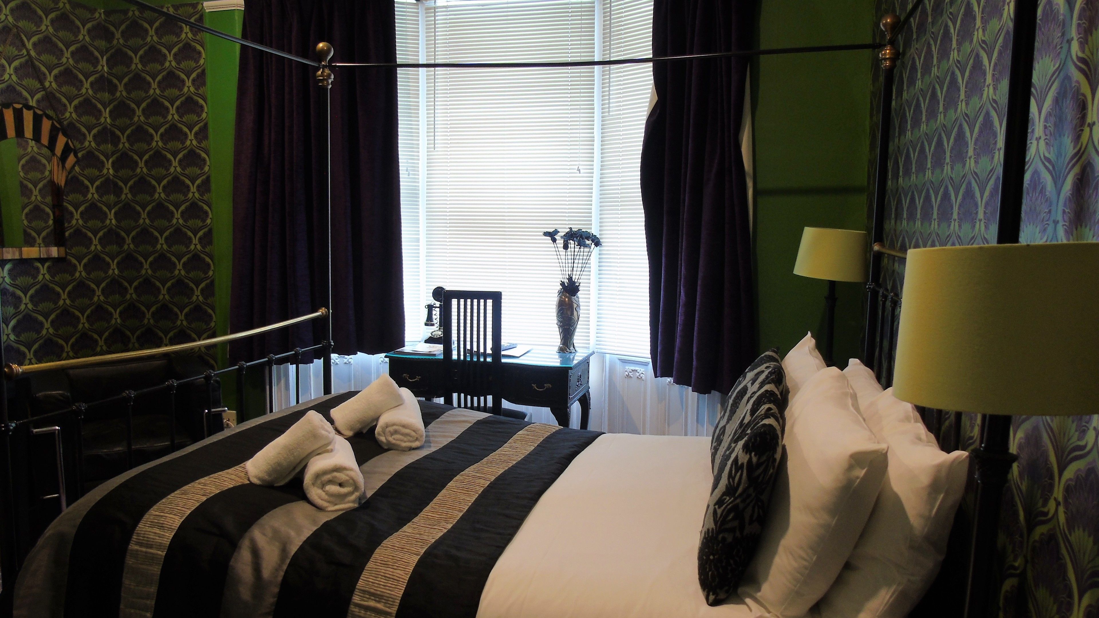 A hotel room with a bed, a desk, a chair, a lamp, and a window with curtains.