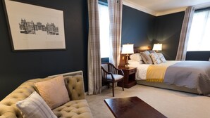 A hotel room with a tufted sofa, a wooden nightstand, a bed with a white comforter and pillows, and a framed picture on the wall. - Duke of Marlborough Hotel (Woodstock)