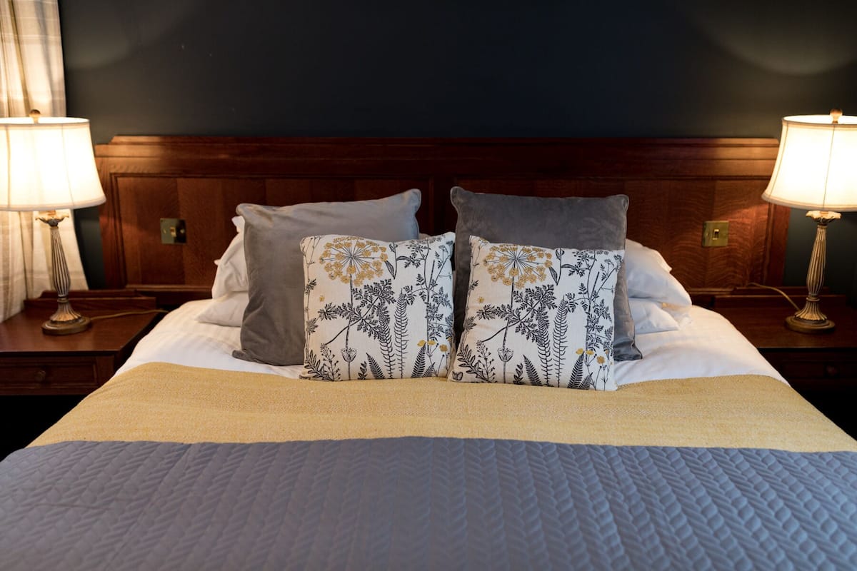 Een netjes opgemaakt bed met een witte dekbedovertrek, twee kussens met bloemmotieven en twee bedlampen op houten nachtkastjes.