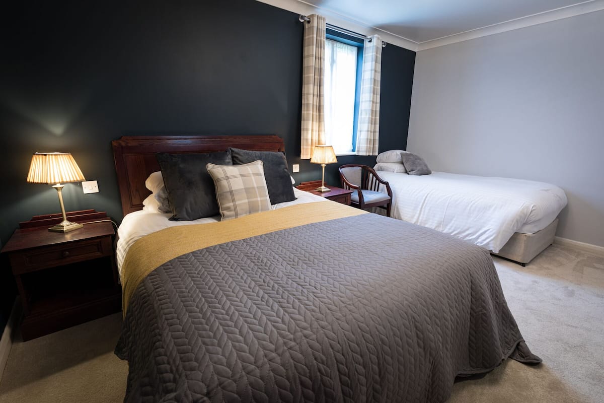 Een hotelkamer met een groot bed, een kleiner bed, twee nachtkastjes met lampen, een stoel en een raam met gordijnen.