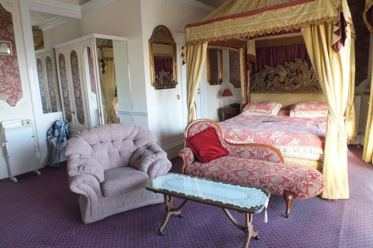 A bedroom with a four-poster bed, a chaise lounge, a vanity with a mirror, and a small table.