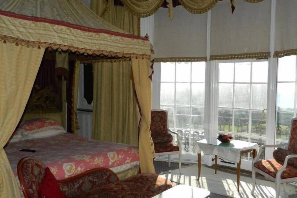 A bedroom with a canopy bed, a sitting area with chairs and a table, and large windows offering a view.
