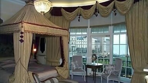 A bedroom with a four-poster bed, a balcony with chairs, and a view of buildings through large windows. - The Prince Regent Hotel (Brighton)
