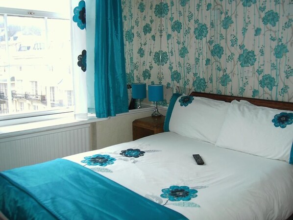 A neatly made bed with a floral bedspread, a remote control on the bed, and a nightstand with two lamps. - The Prince Regent Hotel (Brighton)