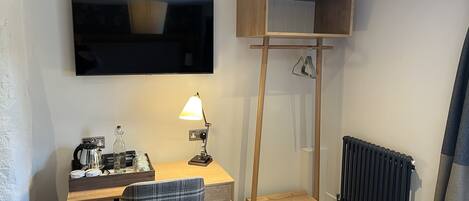 A compact hotel room with a desk, chair, TV, lamp, and a small wooden shelf.