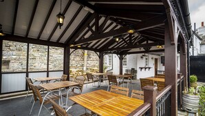 An outdoor patio with wooden tables and chairs, stone walls, and hanging lanterns. - Queens Head Inn (Penrith)