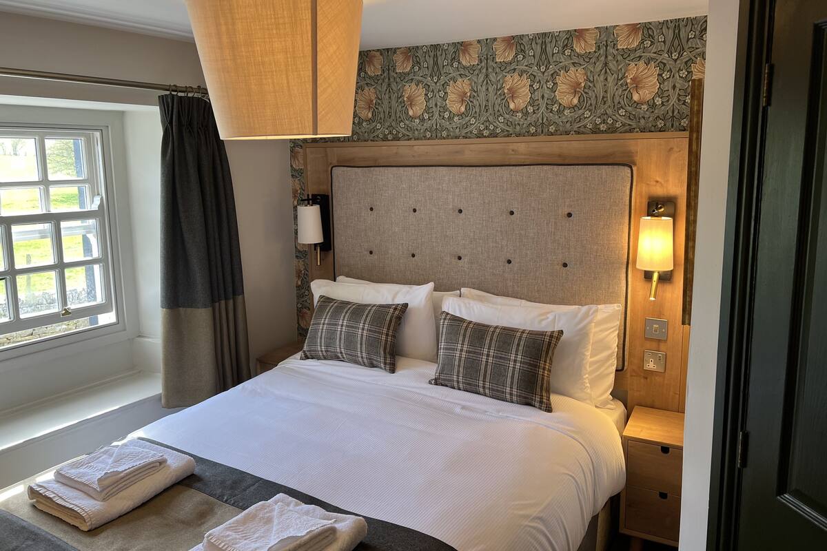 A neatly made bed with a headboard, two bedside lamps, and a window with curtains.