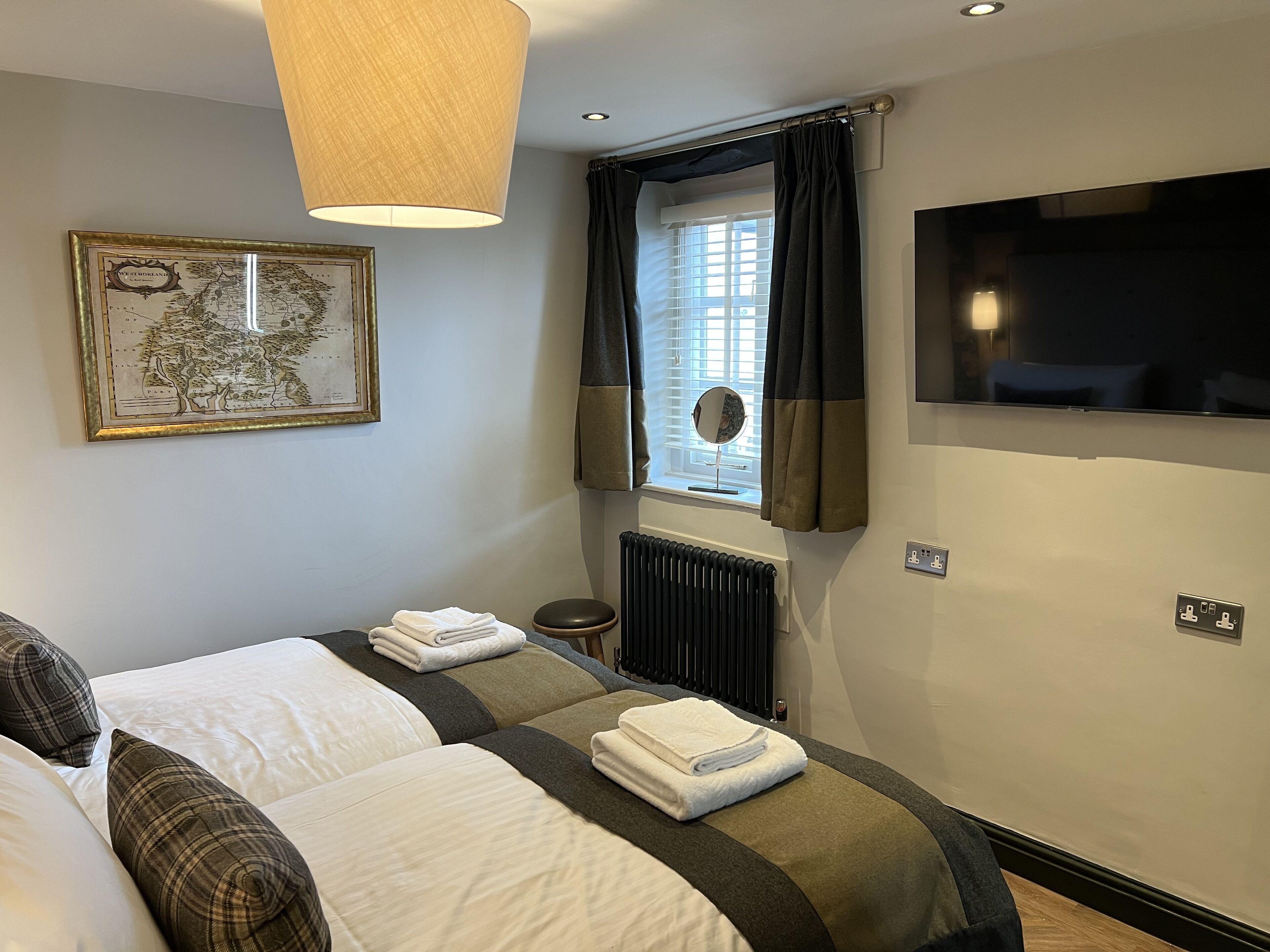 Ein Hotelzimmer mit zwei Betten, einer Karte an der Wand, einem Fenster mit Vorhängen, einem Heizkörper und einem an der Wand befestigten Fernseher.