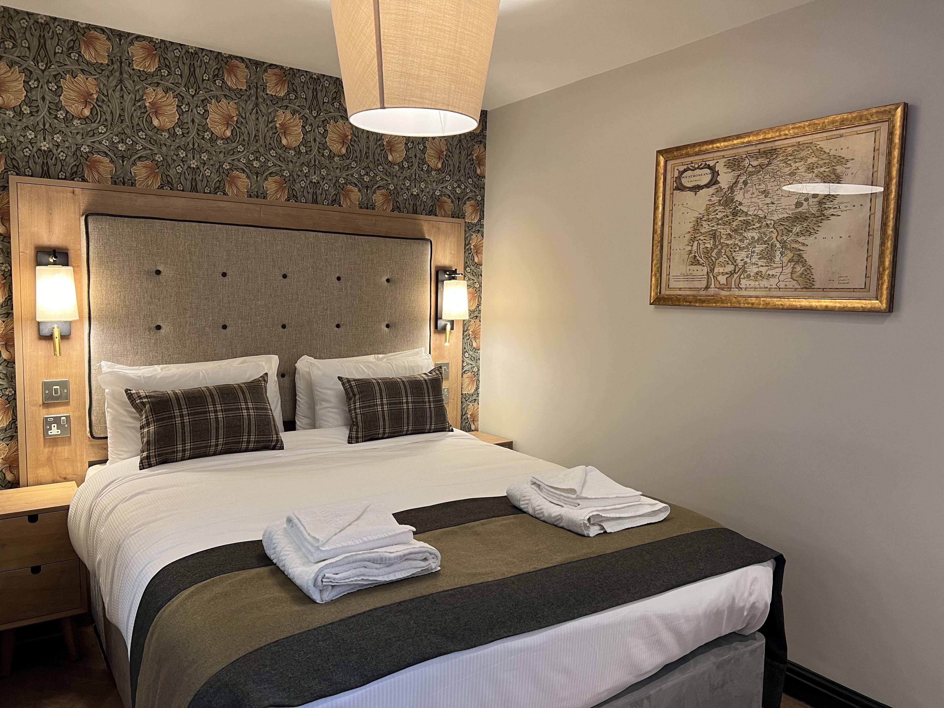 A neatly made bed with white and plaid pillows, a folded towel, and a headboard with wall-mounted lights.