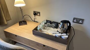 A hotel room with a wooden desk, a lamp, a tray with jars, a coffee pot, and a power outlet.