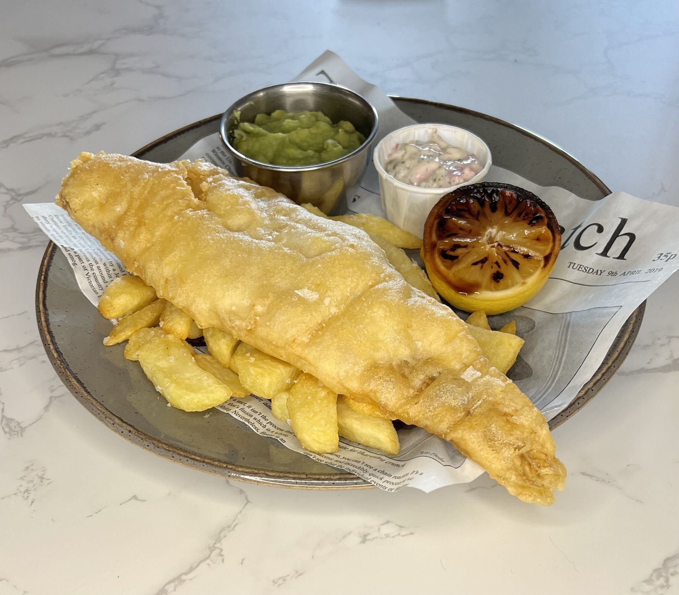 A plate of fish and chips with a side of mushy peas and tartar sauce, served on a newspaper.