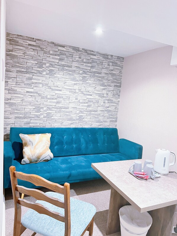 Une pièce avec un canapé bleu, une chaise en bois, une petite table avec une bouilloire et un mur décoré d’un motif imitant la pierre.