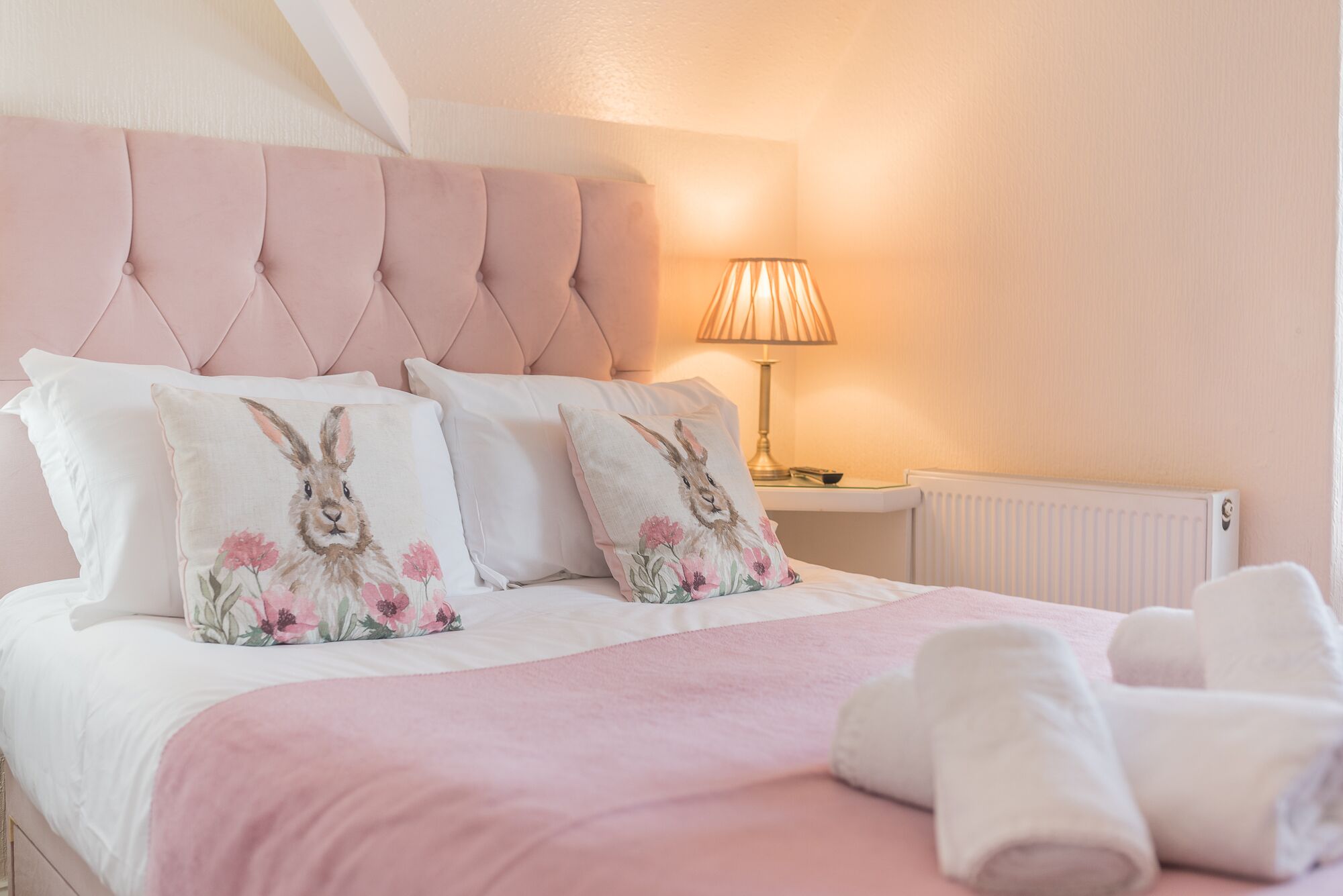 Un lit bien fait, avec une literie rose et des coussins décoratifs, une lampe de chevet et des serviettes enroulées posées sur le sol.