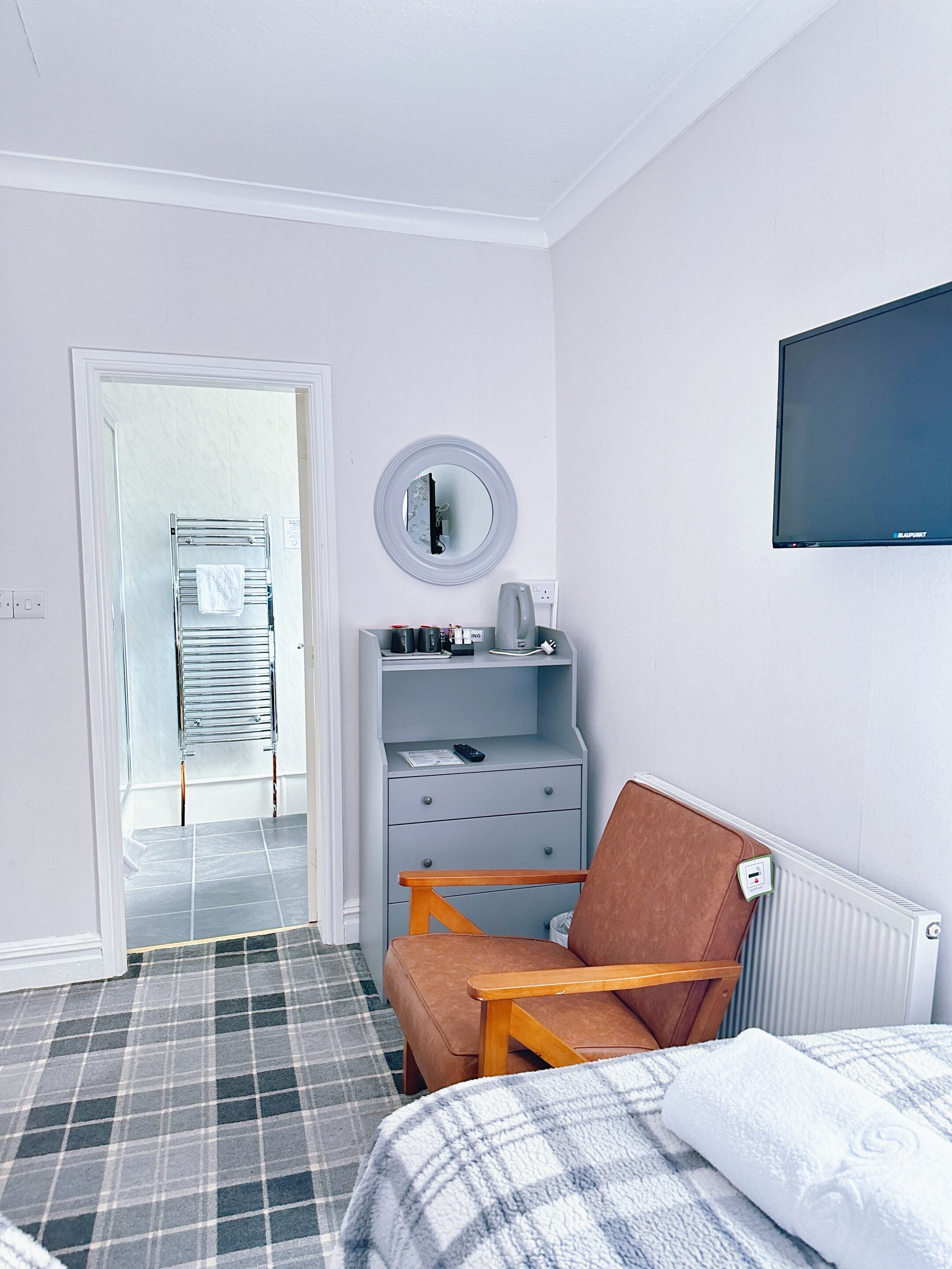 A hotel room with a bed, a chair, a desk with a mirror, a television, and a radiator.