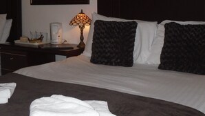 A neatly made bed with white linens, a dark brown throw blanket, and two black pillows. There is a bedside table with a lamp, a coffee maker, and a small tray with cups and a container.