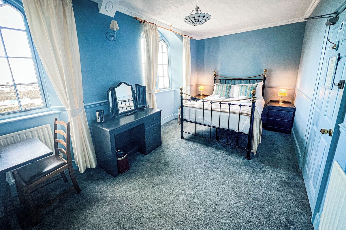 A bedroom with a four-poster bed, a bedside table with a lamp, a dresser, a chair, and a window with curtains.