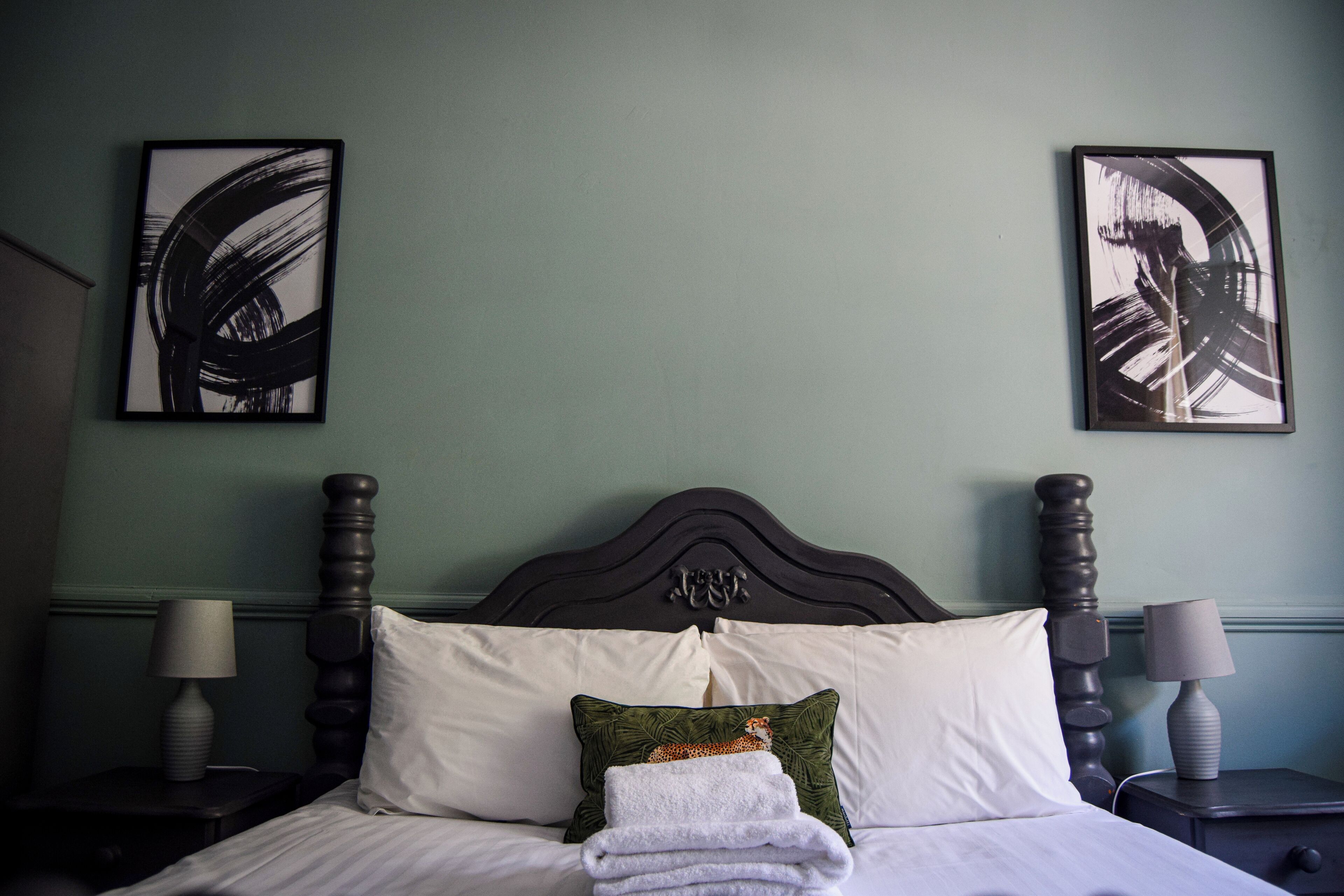 A neatly made bed with white linens, a green pillow, and a folded white towel. There are two bedside tables with lamps, and artwork is displayed on the wall above the bed.