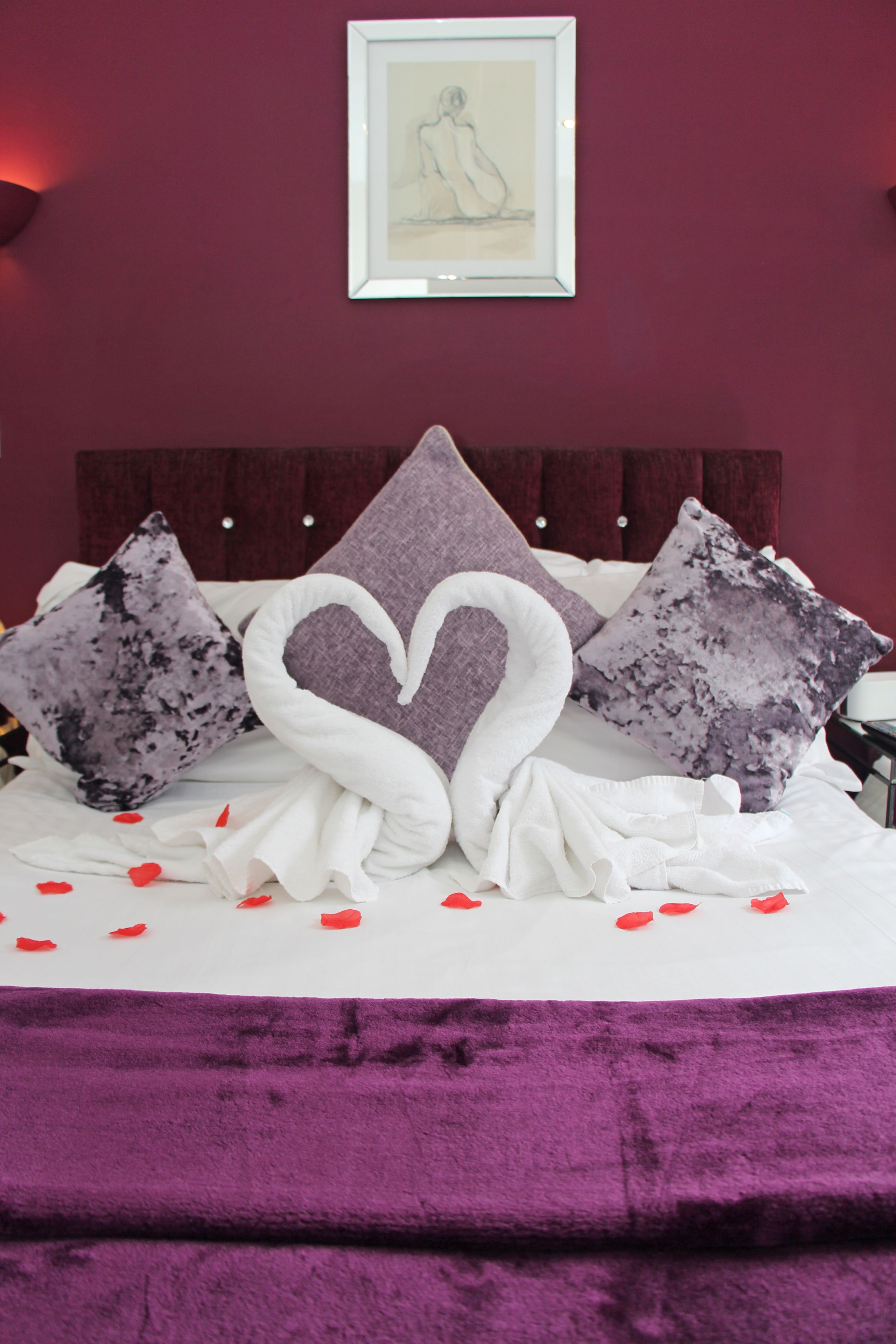 A bed with a heart-shaped pillow, a framed picture on the wall, and rose petals on the bed.