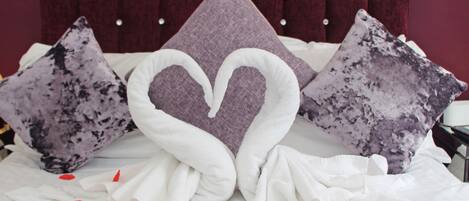 A bed with a heart-shaped pillow, a framed picture on the wall, and rose petals on the bed.