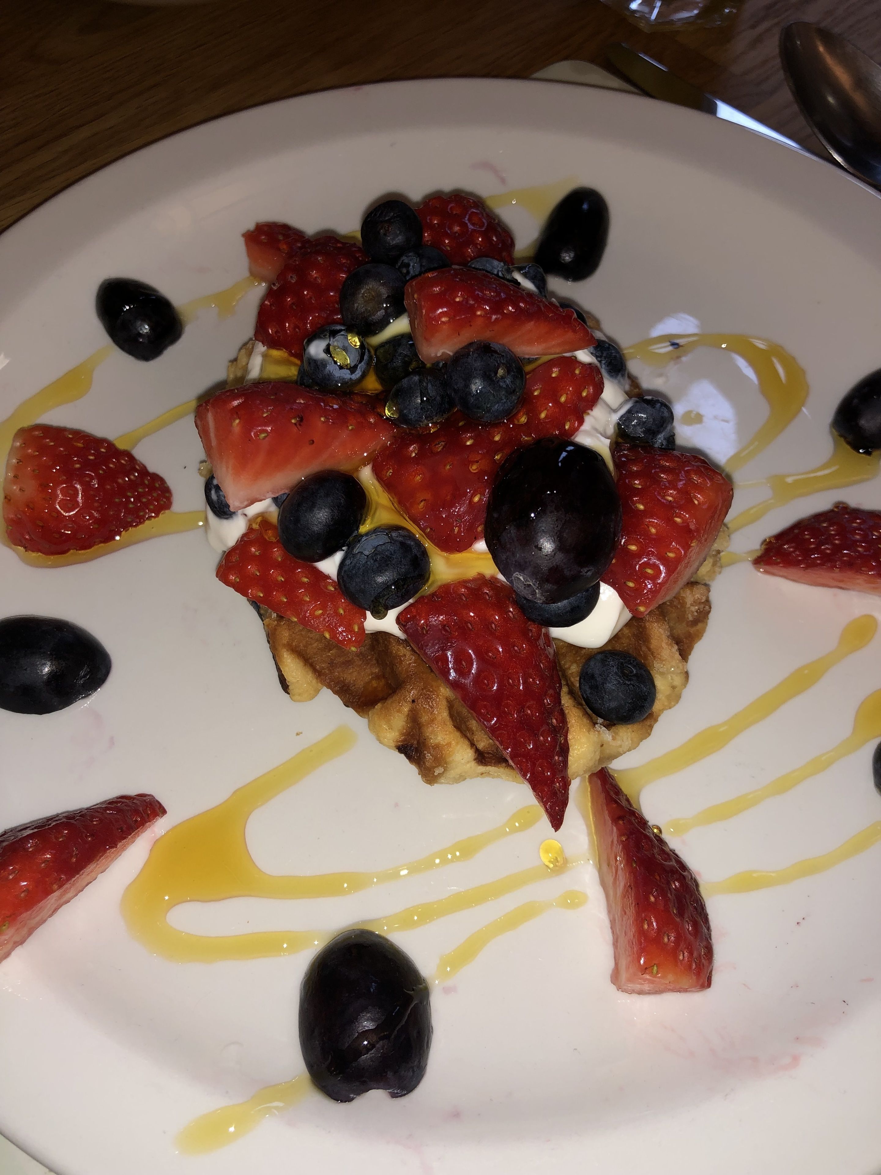Un plato de comida con waffles cubiertos de fresas, arándanos y una cucharada de crema, rociados con jarabe.