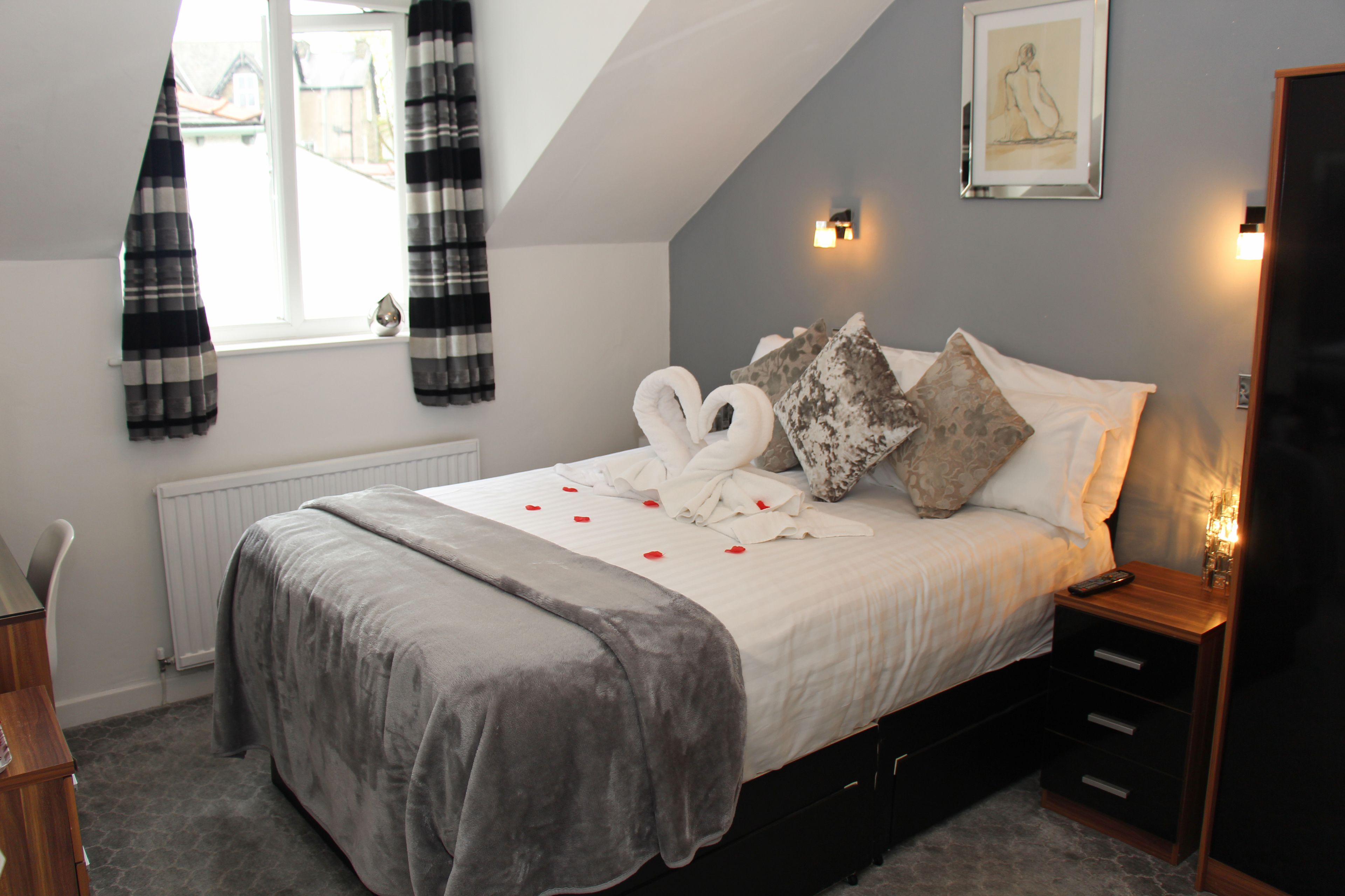 Une chambre à coucher comprenant un lit, une table de chevet, une commode, une fenêtre avec des rideaux et un luminaire fixé au mur.
