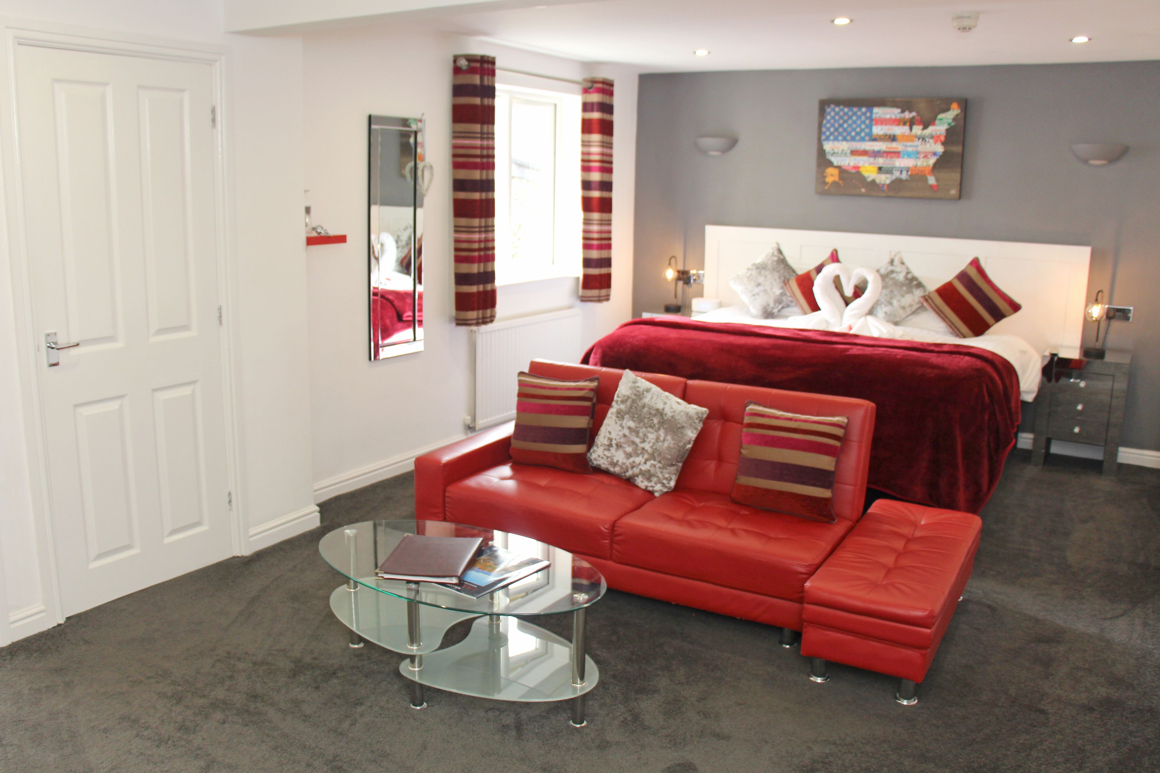 Une chambre d’hôtel moderne avec un canapé en cuir rouge, une table basse en verre et un grand lit recouvert d’une couverture rouge. Il y a une fenêtre avec des rideaux à rayures et une carte accrochée au mur.