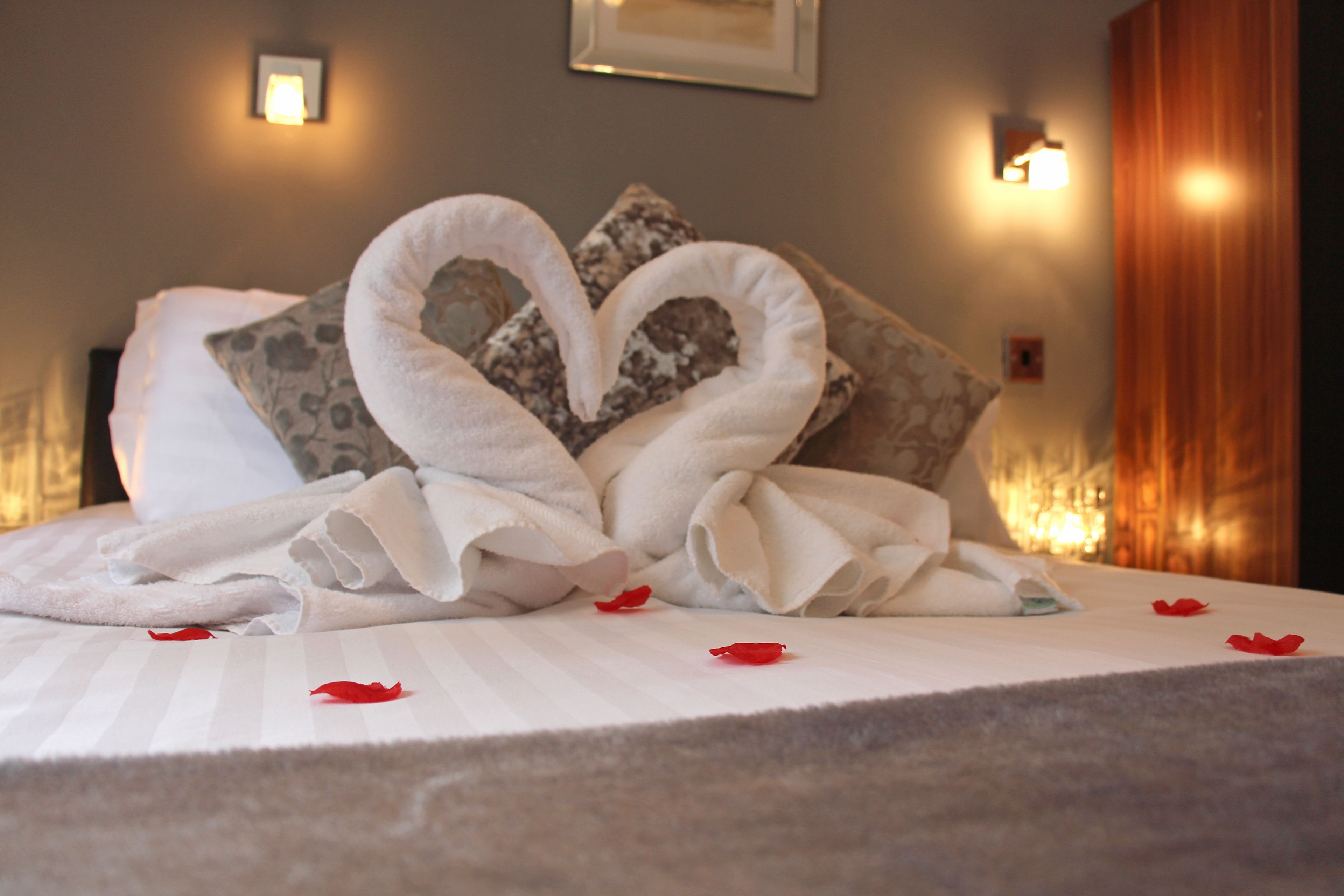 A neatly made bed with a heart-shaped towel arrangement and rose petals on the bedspread.