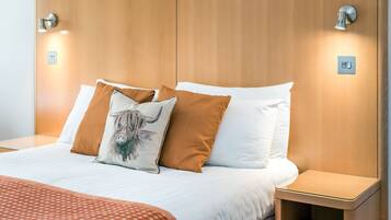 A neatly made bed with white and brown bedding, two wall-mounted light fixtures, and a small bedside table.